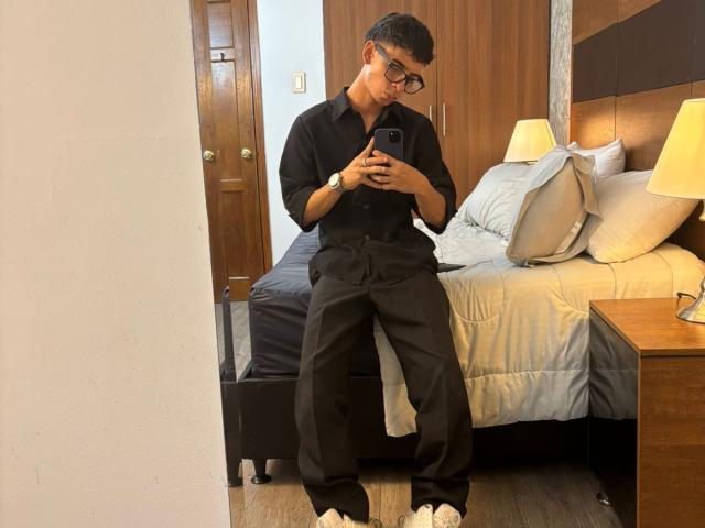 Brian Oconor mit kurzen, schwarzen Haaren und Brille, der ein schwarzes Hemd und eine schwarze Hose trägt und auf einem Bett sitzt und ein Selfie vor einem Spiegel macht, wobei seine weißen Sneakers und seine schwarze Uhr zu sehen sind..