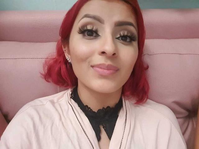 LizBonnet has red hair, wearing a pink shirt, black lace collar, and earrings, with long eyelashes, pink lipstick, and a slight smile. Her tits are not visible.
