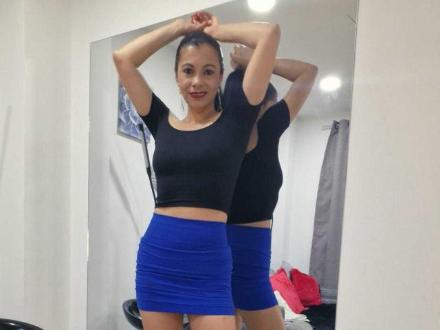 MelissaBaud has small tits, wearing a black crop top and blue skirt, standing in front of a mirror, arms raised, reflecting her. Her arms are tied back.