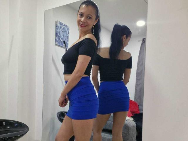 MelissaBaud with dark hair in a ponytail, black off-shoulder top, and blue skirt, showing her ass and tits in a mirror reflection, smiling with red lipstick.