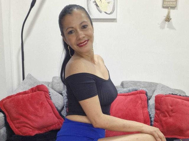 MelissaBaud with black off-shoulder top, blue skirt, red lipstick, and long black hair, smiling, sitting on gray and red pillows, showing her tits and ass.