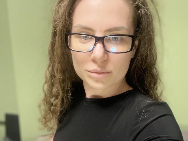 CurlyAmour with curly brown hair, black glasses, and a black shirt, showing a serious face, has a slight pout and visible nose reflection.