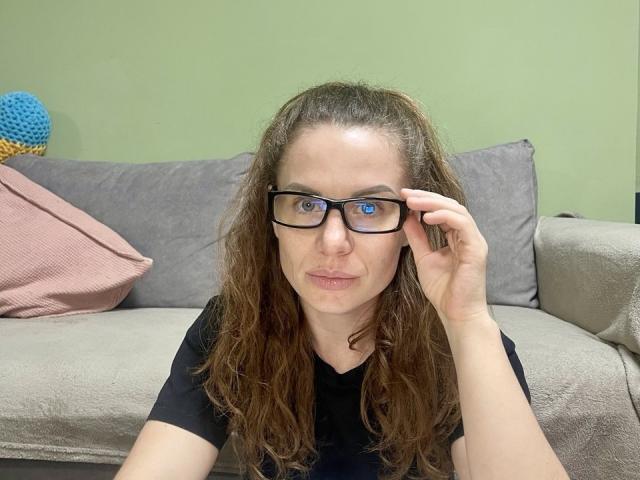CurlyAmour with wavy brown hair, blue eyes, and black glasses, adjusting her glasses with her right hand, sitting on a grey couch, wearing a black shirt.
