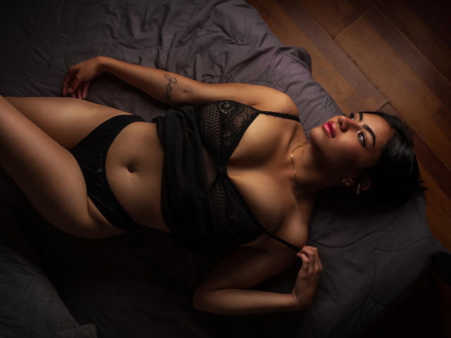 ValeryJhons lying on a bed, wearing black lace lingerie, her tits visible, black panties, tattoo on her right arm, dark hair, red lipstick, wooden floor, dark gray bedsheet.
