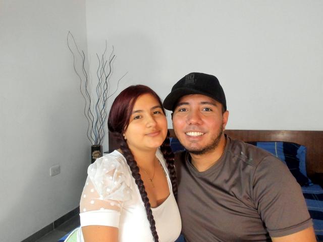 TifannyLov with dark red braids, white polka-dot top, and medium tits, sitting next to a Latino dude in a brown shirt and black cap, smiling.