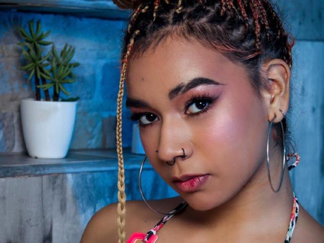 MadissonGreey has braided hair with red tips, hoop earrings, nose ring, pink eyelids, and a serious expression. She's wearing a floral halter top, showing her tits slightly.