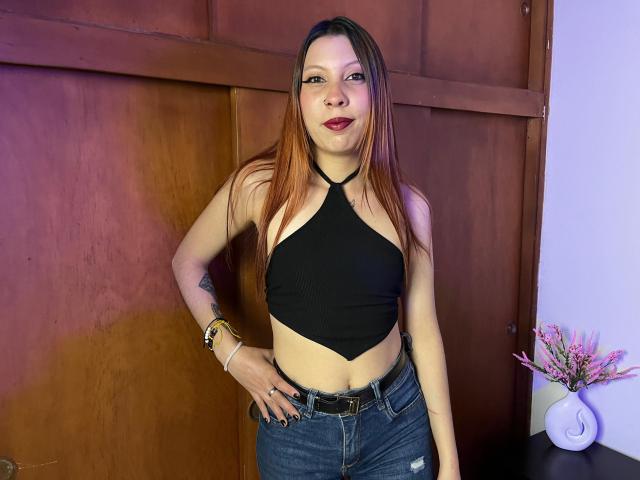 MelanyDurango with long brown hair, black halter top, small boobs, blue jeans, and a belt, standing, hand on hip, smiling, wearing red lipstick, wooden background.