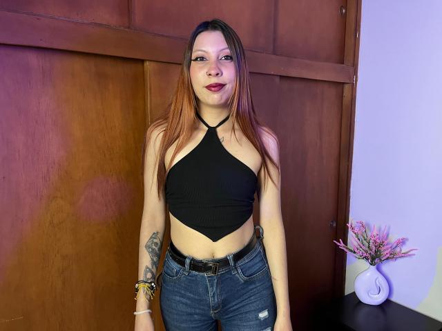 MelanyDurango with long brown hair, black halter top showing her small tits, and blue jeans, standing against a wooden wall, smiling with red lipstick, tattoo on right arm.
