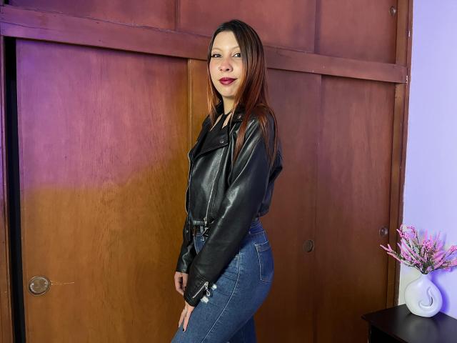 MelanyDurango with long brown hair, wearing a black leather jacket and high-waisted blue jeans, standing with a smile, showing her ass, against a wooden door.