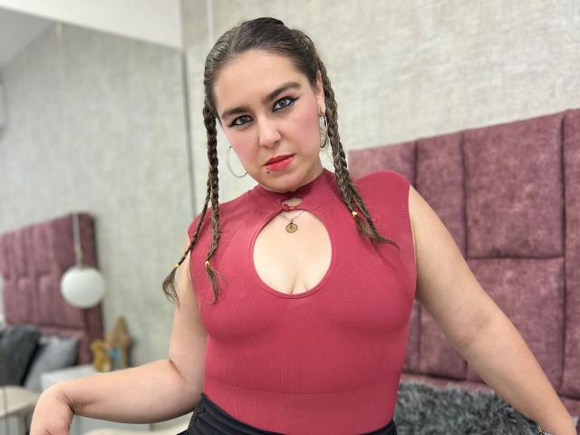 TaylorNova, avec ses cheveux bruns tressés, son rouge à lèvres rouge et un haut rouge moulant sans manches qui révèle ses petites seins et une découpe. Elle regarde l'appareil photo avec un léger sourire..