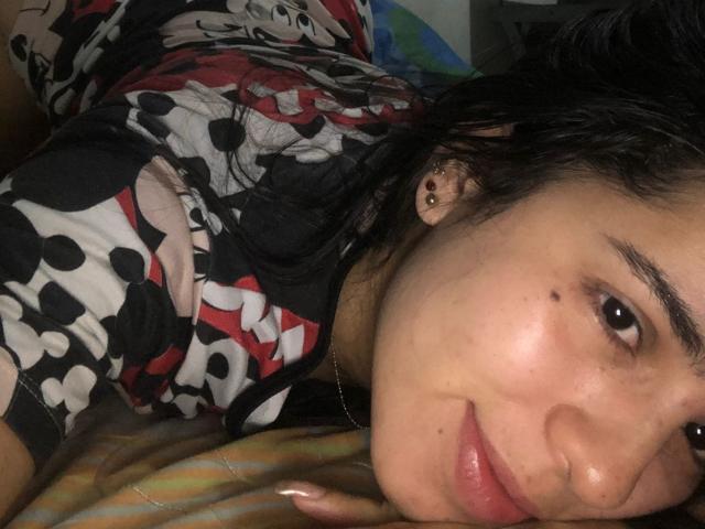 AlanaReyes lying on a bed, face up, smiling, wearing a black, red, and white patterned shirt, showing a small beauty mark on her cheek, with earrings.