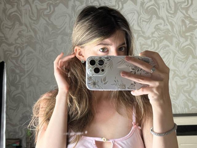 VinnaSkyHott with long blonde hair, green eyes, holding a phone with a leaf pattern case, wearing a pink, lace-trimmed top, showing her tits in a mirror selfie.