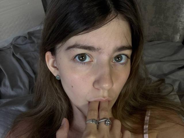 VinnaSkyHott with brown eyes, long brown hair, sucking her finger with a silver ring on it, wearing a pink top, and a small mole on her right cheek.