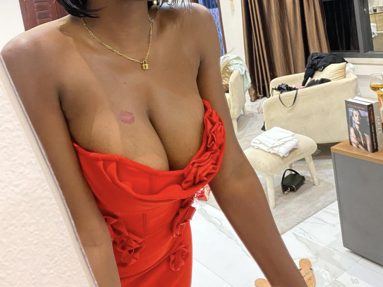 SexyAkena with large, round tits in a red, ruffled dress, showing a kiss tattoo on her chest, wearing a gold necklace, in a modern, well-lit room.
