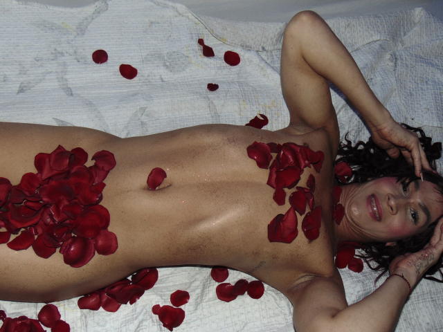 SarithaCute está desnuda sobre una cama blanca, cubierta de pétalos de rosa roja, con las manos en su cabello rizado y oscuro, mostrando sus pechos y vulva, con una sonrisa relajada..
