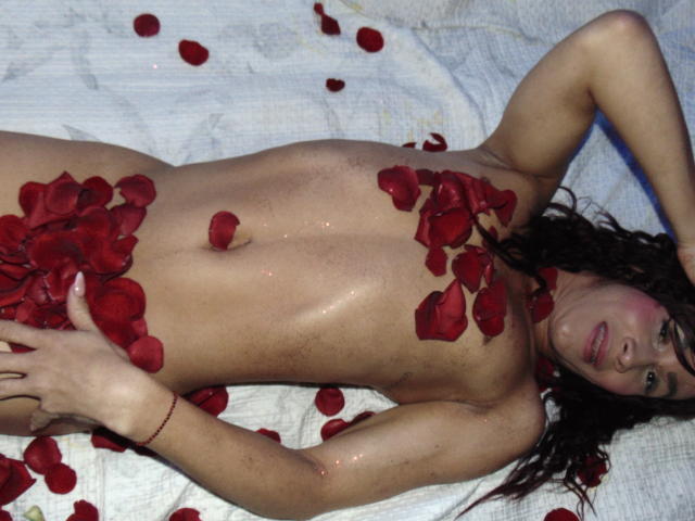 Saritha, nackt, liegt auf einem weißen Handtuch, bedeckt mit roten Rosenblättern, mit Blättern auf Brüsten, Bauch und Vulva, Arme über dem Kopf, lockiges braunes Haar, leicht feucht, entspannter Gesichtsausdruck..