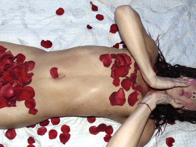 SarithaCute, naked, lying on white bed with red rose petals covering her tits and pussy, arms above head, long dark wavy hair, pink nails, tan skin, slight tan lines.