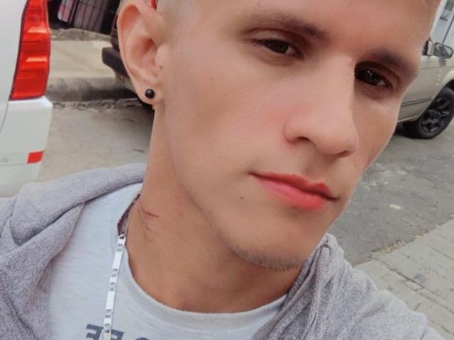 LiamStone has a shaved head, black ear studs, light skin, and a small scar on his neck. He's wearing a gray hoodie and a white shirt with a necklace.