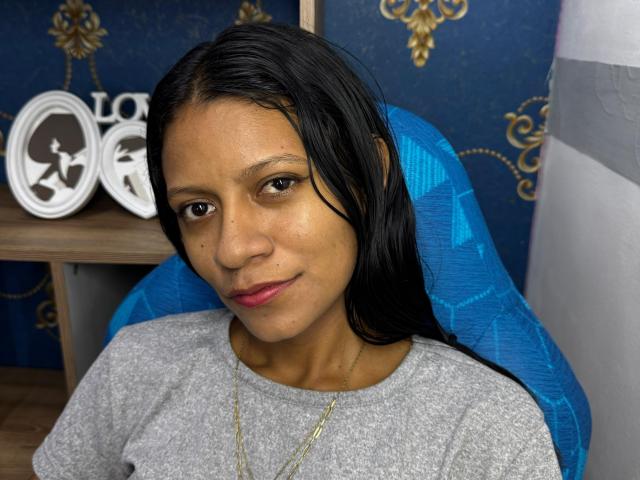 MonnicaBella has brown skin, black straight hair, and brown eyes, wearing a gray shirt and gold necklace, sitting in a blue chair, with a small mole on her left cheek.