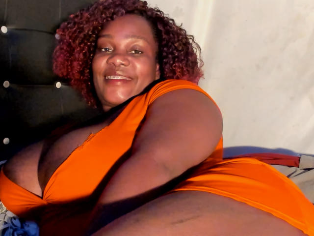 AgathaBlossom, a curvy black woman with red curly hair, wearing an orange dress, lying on a bed, smiling at the camera, showcasing her large boobs.