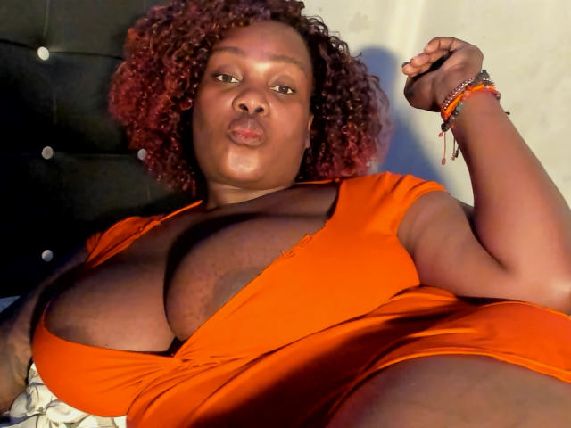 AgathaBlossom with curly red hair, wearing an orange dress, showing large tits, pouty lips, right arm raised, bracelets on wrist, lying down, smiling, dark skin, bedroom.