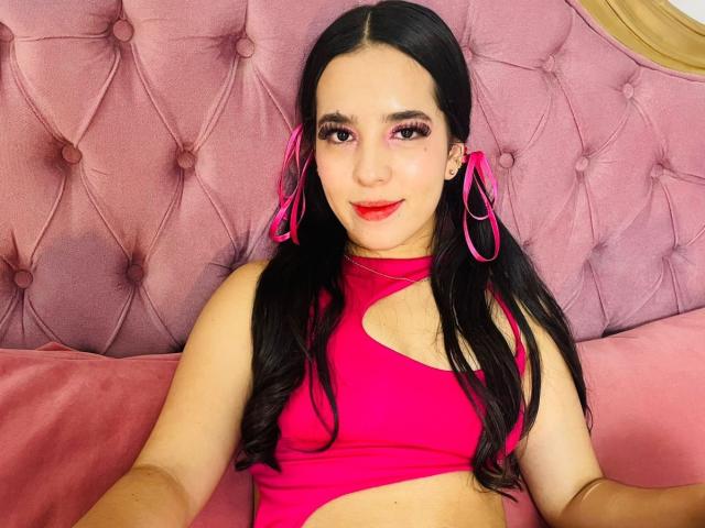 AlanaReyes with long black hair, pink ribbons, and red lipstick, wearing a hot pink cutout top, sitting on a pink tufted couch, showing her tits.