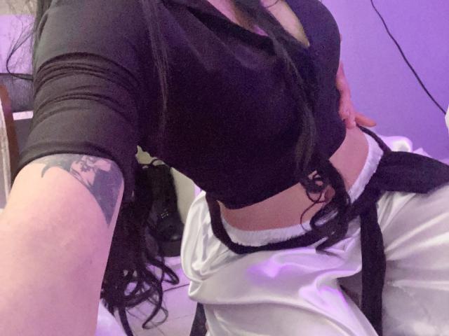 una gatta con il petto nero in evidenza, pantaloni bianchi, cintura nera, tatuaggio sul braccio destro, capelli neri, ventre scoperto, sfondo viola, leggermente piegata, che si fa un selfie..