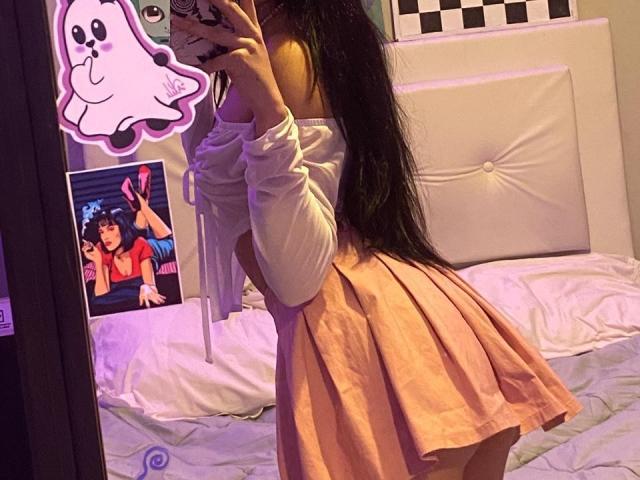 Een vrouw met lang zwart haar, een witte top met open schouders en een roze plooirok, die haar achterwerk laat zien in een spiegelzelfportret. De reflectie bevat een sticker van een spook en een foto..
