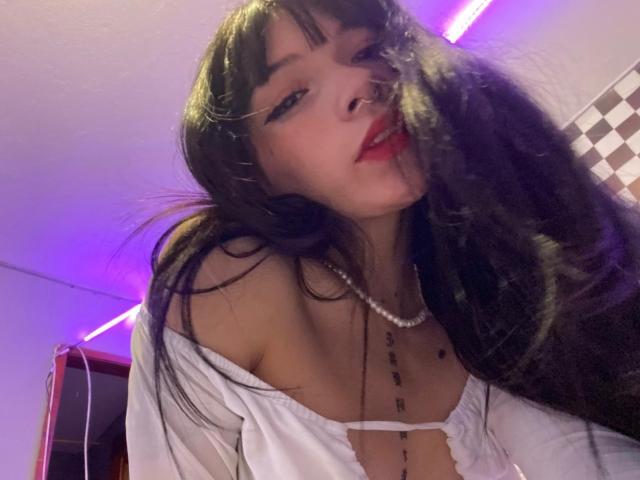 una gatta nera con lunghi capelli neri, rossetto rosso, piercing al naso, top bianco con una spalla scoperta, collana di perle, che guarda in basso, con la testa leggermente inclinata, luce al neon viola sullo sfondo, sorriso appena accennato..
