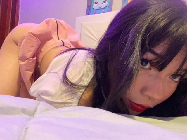 Una ragazza con capelli neri, rossetto rosso e piercing al naso, seduta su un letto, indossa una gonna rosa e una camicia bianca, con i glutei in evidenza, guarda verso la fotocamera, illuminazione viola, cuscino bianco in primo piano..