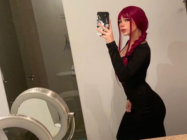 Una ragazza con capelli rossi raccolti in una treccia, che indossa un vestito nero, sta scattando un selfie allo specchio, mostrando il sedere e i piccoli seni, e tiene in mano un telefono con una custodia nera..