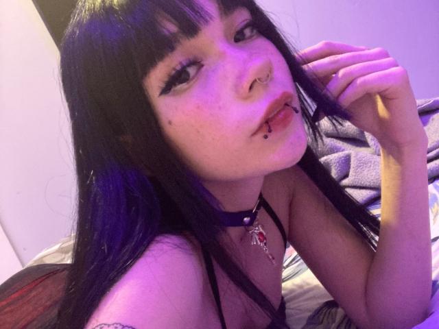 una gatta di nome Nina, con capelli neri e riflessi viola, un anello al naso, un piercing al labbro, lentiggini, che indossa una collana nera, sdraiata sul letto, illuminazione viola, che guarda la fotocamera, con una mano vicino al viso..