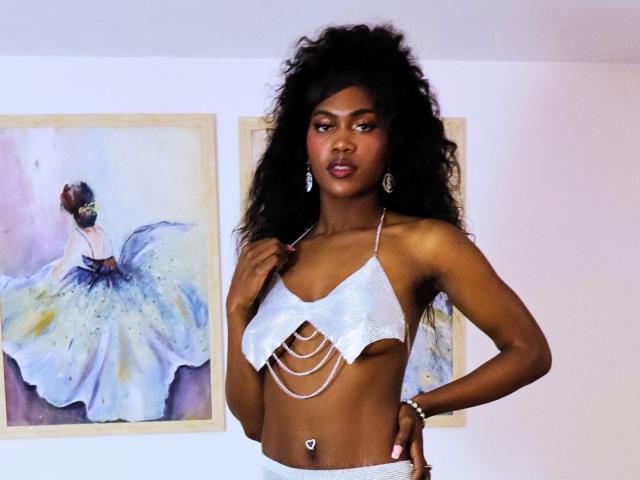 MeganMonsalve with dark curly hair, wearing a shiny white halter top with cut-out rings, showing her small tits and a heart-shaped belly button. Two watercolor paintings in background.