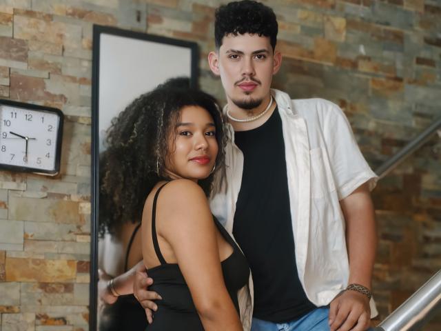 AshleyAndJack, avec des cheveux bouclés, un débardeur noir laissant entrevoir ses seins, se tient à côté de Jack, qui a les cheveux courts et bouclés, porte une chemise blanche et un t-shirt noir, et a une main sur ses fesses. L'arrière-plan est un mur de pierre..