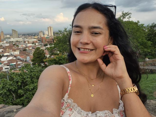 LilianaPink with medium tits, wearing a floral lace top, smiling, dark hair, gold necklace, watch, and sunglasses on head, holding her cheek in a selfie.