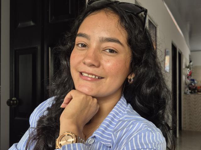 LilianaPink with curly black hair, wearing a blue striped shirt, smiling with her chin on her hand, brown eyes, gold watch, black sunglasses on head, subtle makeup, earrings.