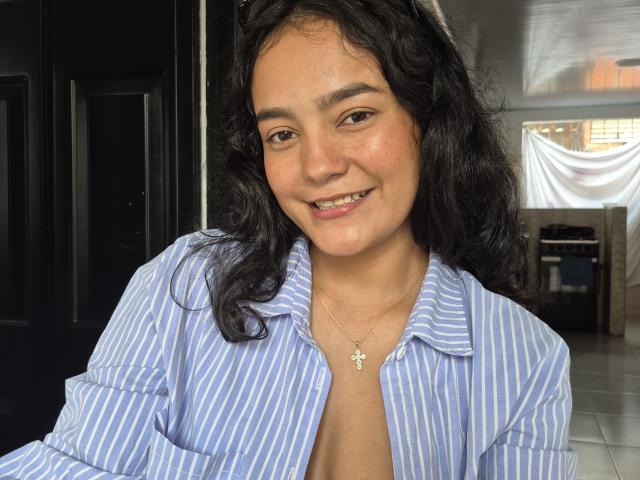 LilianaPink has curly black hair, a cute smile, and is wearing a blue and white striped shirt, with a small, visible cleavage and a cross necklace.