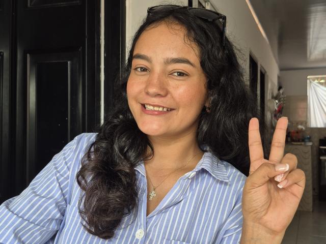LilianaPink with curly black hair, blue striped shirt, smiling, showing a peace sign, has small tits, tan skin, black eyewear on head, necklace with star pendant, indoors.