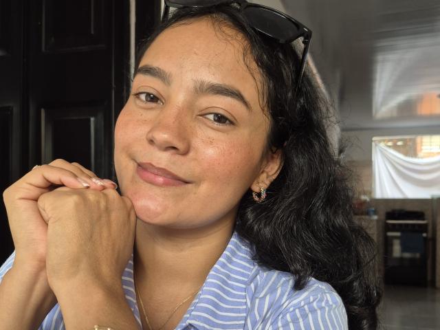 LilianaPink has medium brown skin, black wavy hair, brown eyes, small nose, and subtle smile. She's wearing a blue-striped shirt, small earrings, and black sunglasses on her head.