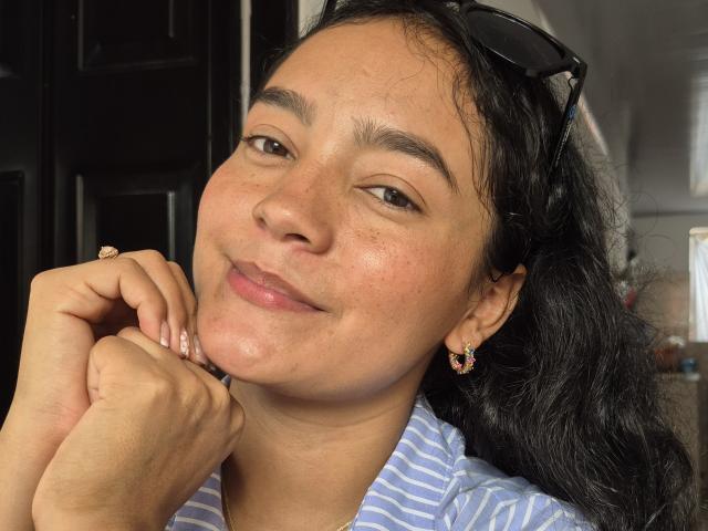 LilianaPink has medium brown skin, black wavy hair, and is smiling with her hand near her chin. She's wearing a striped shirt, earrings, and has glasses on her head.