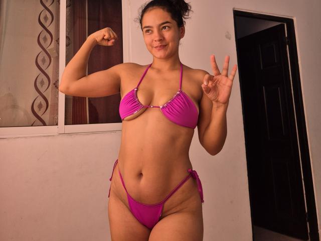 LilianaPink in a pink bikini, flexing her right arm, showing a peace sign with her left hand, smiling with medium tits and a slightly chubby, curvy ass.