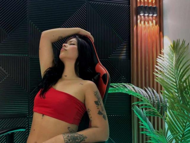 AbbyPons with black hair, red tube top, tattoos, arm raised, pouty expression, sitting on red chair, green plant on right, wood and black panel background.
