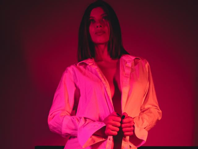BeverlyBonnet with pink-red lit face, wearing an unbuttoned white shirt, exposing her tits, holding the shirt, straight brown hair, looking up, red glow, hands on shirt.