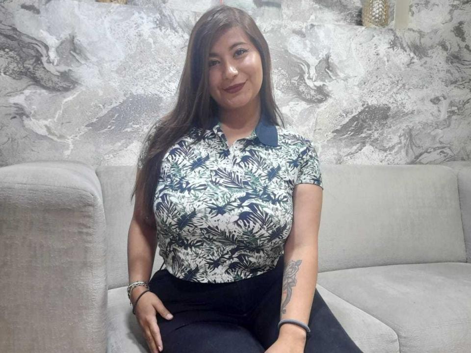 VeroniicaJhonson with large tits, wearing a green leaf pattern shirt and black pants, sitting on a gray couch, smiling, long brown hair, tattoo on right forearm.