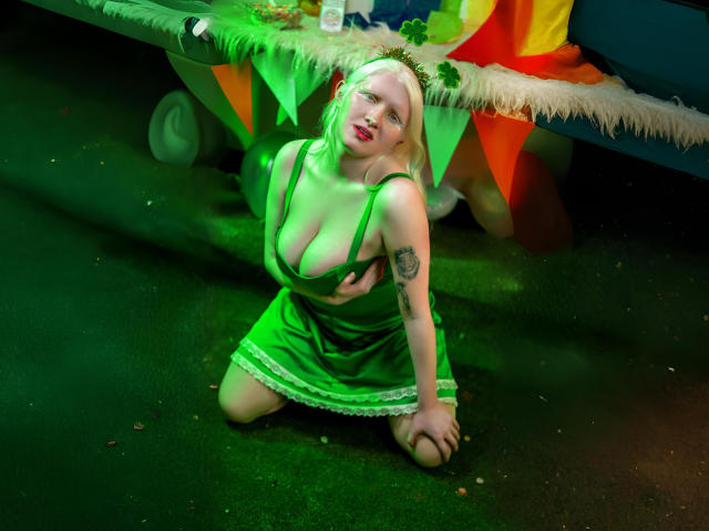 SilverLady with green dress, kneeling on grass, green hair, green headband, large tits, tattoo on right arm, looking up, under a table with green, yellow, and orange decorations.