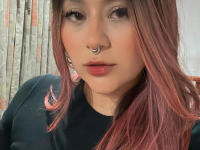 LorraineV has pinkish-red hair, a silver nose ring, and red lipstick. She's wearing a black shirt with pink accents. Her eyes are slightly open, looking at the camera.