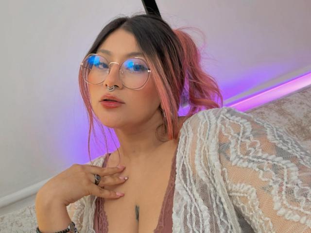 LorraineV with pink-tipped hair, glasses reflecting blue, nose ring, pink lips, lace top, hand on chest, subtle tattoo, purple lighting, looking at camera.