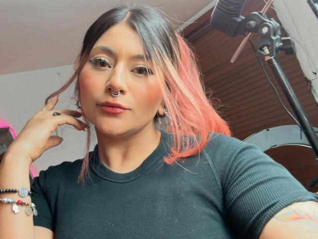 LorraineV with pink-dyed hair, nose ring, black tee, and tattoos, looking at camera, hand near face, camera light in background.