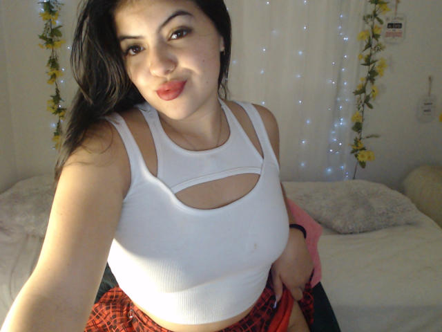 AlissonS with dark hair, pouty red lips, white cutout tank top, small tits, red plaid skirt, holding her skirt up, sitting on bed, soft white lights in background.