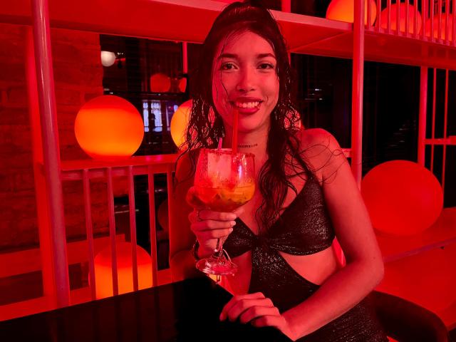 KrystalEvans with wet black hair, holding a cocktail with a straw, smiling, wearing a black glittery bra, sitting in a red-lit bar. Her tits are visible.